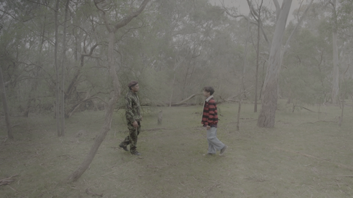 Soldier in camouflage and a young person in a red-striped jacket stand facing each other in a misty bush clearing.