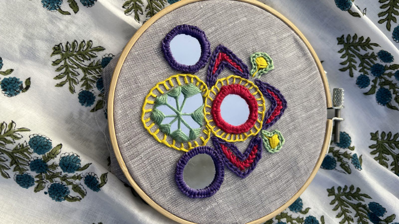 An embroidery hoop with colourful patterns and mirrors stitched to the fabric.