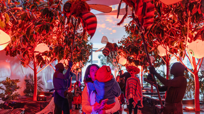 A parent and child walk through a lane flagged in trees and puppeteers moving giant bees on poles.