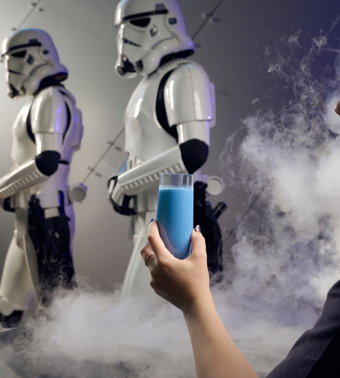 Woman holds blue milk in a smokey room with two stormtroopers in the background.