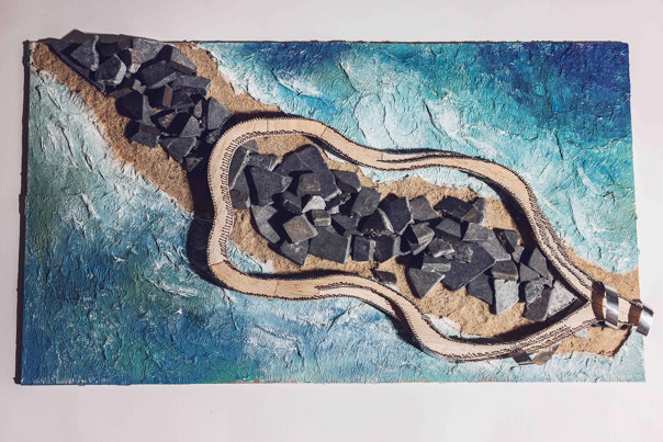 Overhead view of an architectural model showing curved coastal walkway with textured beige surface winding around dark, rocky formations, surrounded by blue ocean with white wave-texture detail.