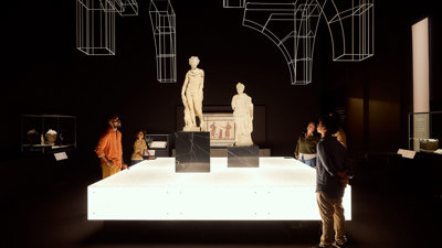 Visitors gathered around two Roman statues on raised plinths within a dramatic, dark exhibition space in ROME: Empire, Power, People exhibition in Melbourne Museum.. 