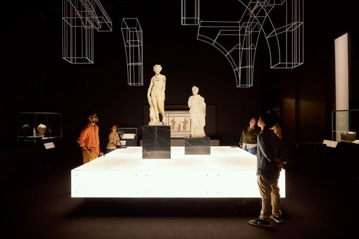 Visitors gathered around two Roman statues on raised plinths within a dramatic, dark exhibition space in ROME: Empire, Power, People exhibition in Melbourne Museum.. 