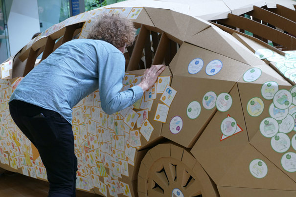 A woman looks in the side window of a life-size cardboard car.