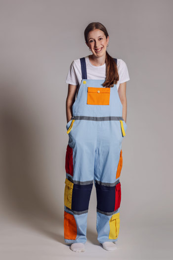 Model wearing light blue overalls with colourful pockets in orange, yellow, and red, featuring reflective grey stripes and navy blue panels at the knees, paired with a white t-shirt.