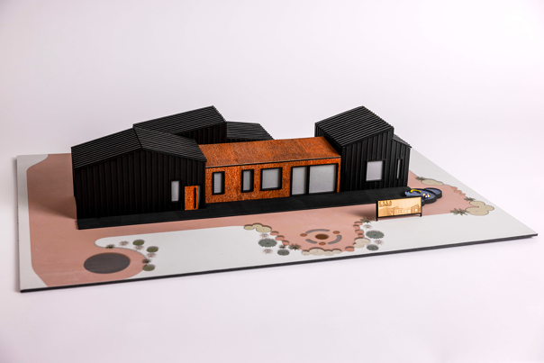 Architectural model of a house with black corrugated roofing, burnt orange walls, and a landscaped base.