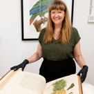 Woman smiling at the camera, holding a rare book open in front of her. She is wearing blue gloves