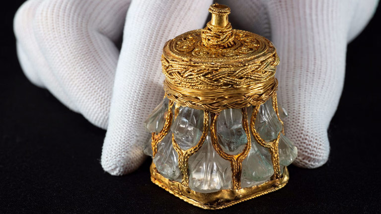 A close-up image of a hand, wearing a white glove, holding a rock crystal jar. This rock crystal jar is one of the treasures of the Galloway Hoard. It is a small, delicate item carved from crystal and decorated with gold.