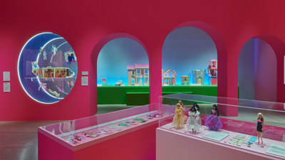  An image of a room with large pink wall with arched doorways and a circular window. In the background you can see a selection a Barbie Dreamhouses. In the foreground you can see two display cases with Barbie accessories and various Barbie dolls.    